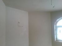 Wnętrze pomieszczenia po nałożeniu świeżego, jasnego tynku. Widoczne gładkie ściany i sufit, zarys okna w tle, oraz drobne niedoskonałości w strukturze tynku na ścianie po lewej stronie.